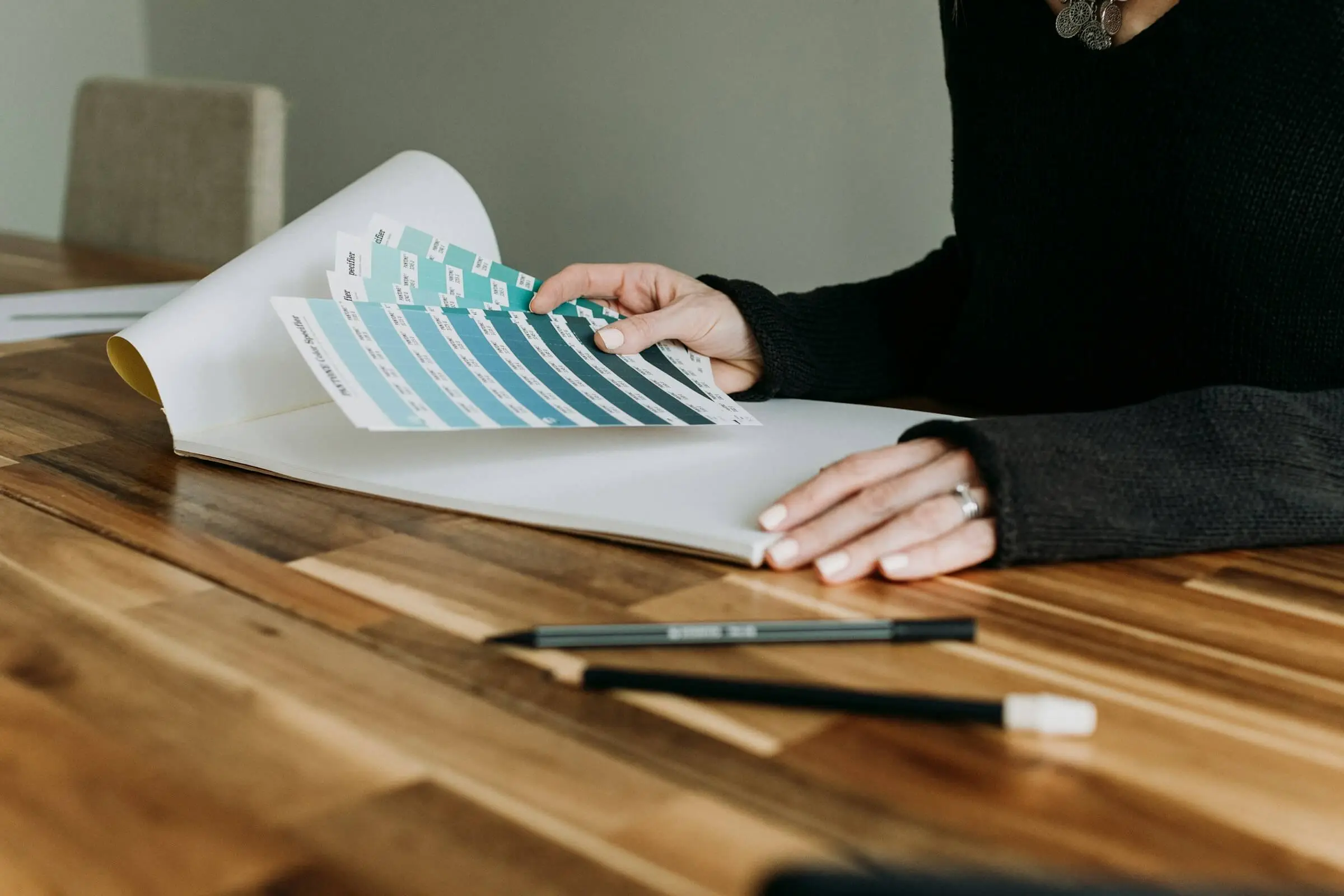 A designer compares teal color swatches over an open sketchbook on a wooden table.